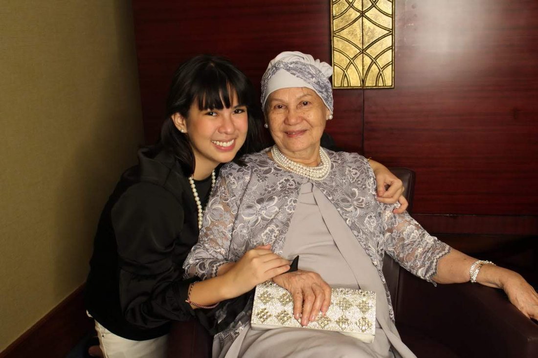 Paulina and her grandmother, older, seated together and smiling, her grandmother in a headwrap holding a beaded clutch.