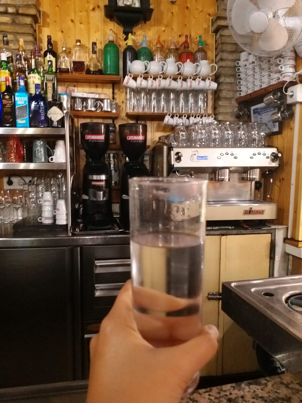 A hand holding up a glass at an old-school Spanish bar, rows of coffee grinders and siphons behind the counter.