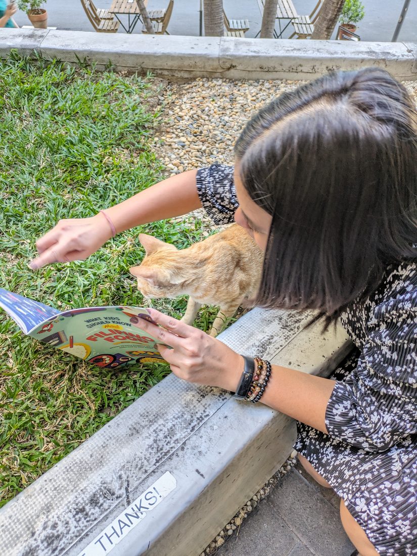Paulina reaching toward a street cat while holding open a children's book about Filipino foods.