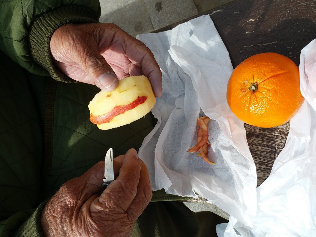 An elderly man's hands peeling an apple with a small knife, an orange resting nearby on a wooden bench.