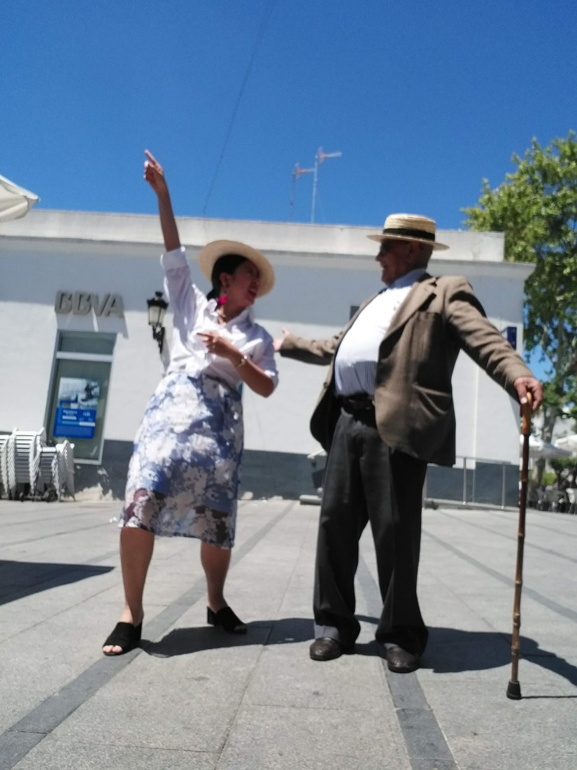 Paulina and Santiago dancing together in a plaza, him in a straw boater hat with a cane, her pointing up at the sky.